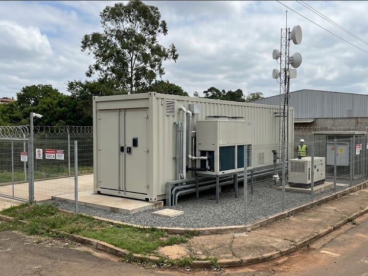 Edge data center in a beige container with cooling units, enclosed by a fence with razor wire and warning signs, next to an antenna tower