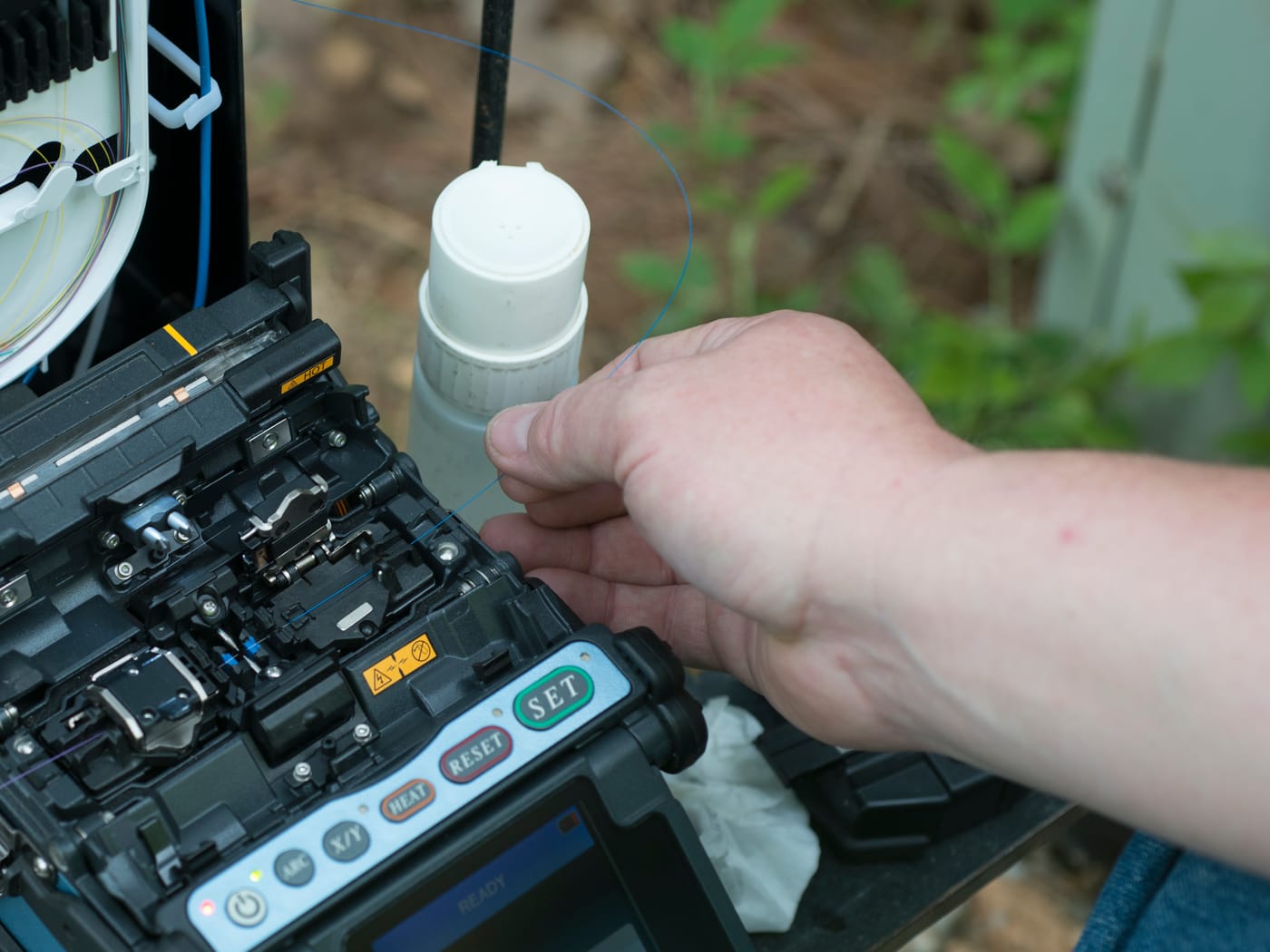A technician's hands splicing optical fiber with a fusion splicer, next to an open splice closure
